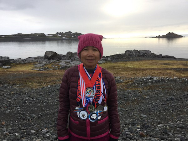 Photo of an older Southeast Asian woman wearing a pink beanie-style hat with cat ears. She has multiple medals around her neck and is standing in front of a body of water outside.